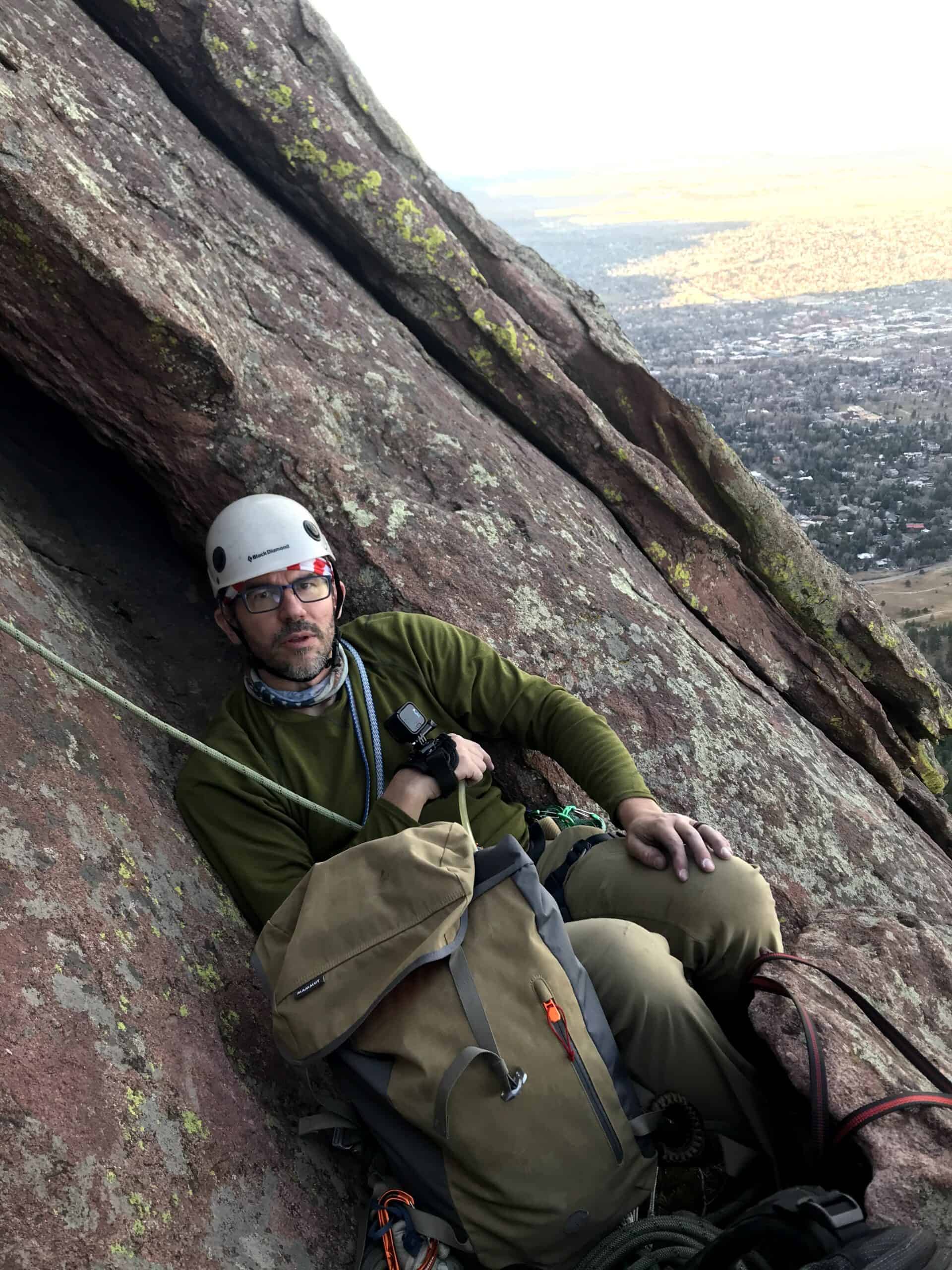 Matt in a helmet and green jacket resting on a steep rock face, with the plains far below